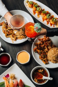 Two signature cocktails being toasted above dishes of sushi rolls, hibachi chicken with vegetables and fried rice, assorted nigiri sushi, onion soup, and dipping sauces visible on table.