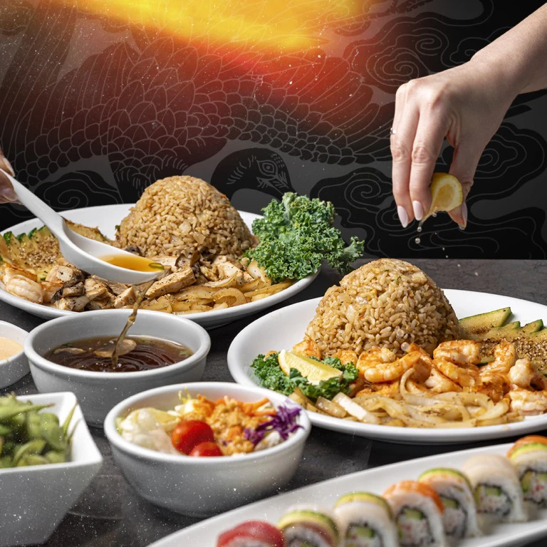 An assortment of Benihana dishes with hibachi rice, shrimp, chicken, sushi rolls, and edamame; hand squeezing lemon over rice.