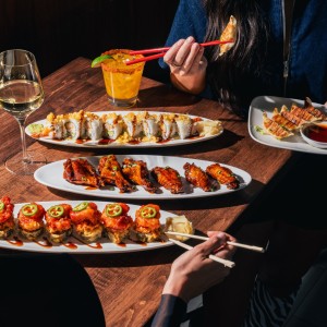 Four sharing plates of Benihana happy hour dishes-sushi rolls, wings, and gyoza-with a guest lifting gyoza with chopsticks. Glasses of wine and a margarita are also on the table.
