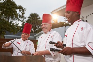 Smiling Benihana chefs in white uniforms and red hats demonstrate teppanyaki skills at an outdoor cooking station.