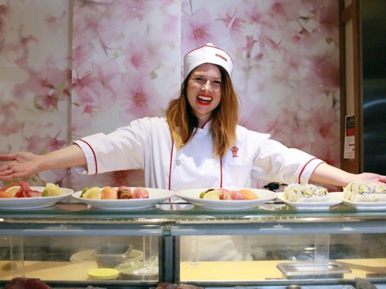 Person dressed in Benihana chef clothing, smiling behind the counter with plates of nigiri, sashimi and sushi rolls ready to serve
