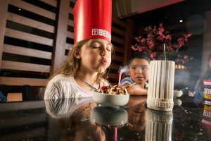 A child wearing a chefs hat blowing out a candle on an ice cream sundae